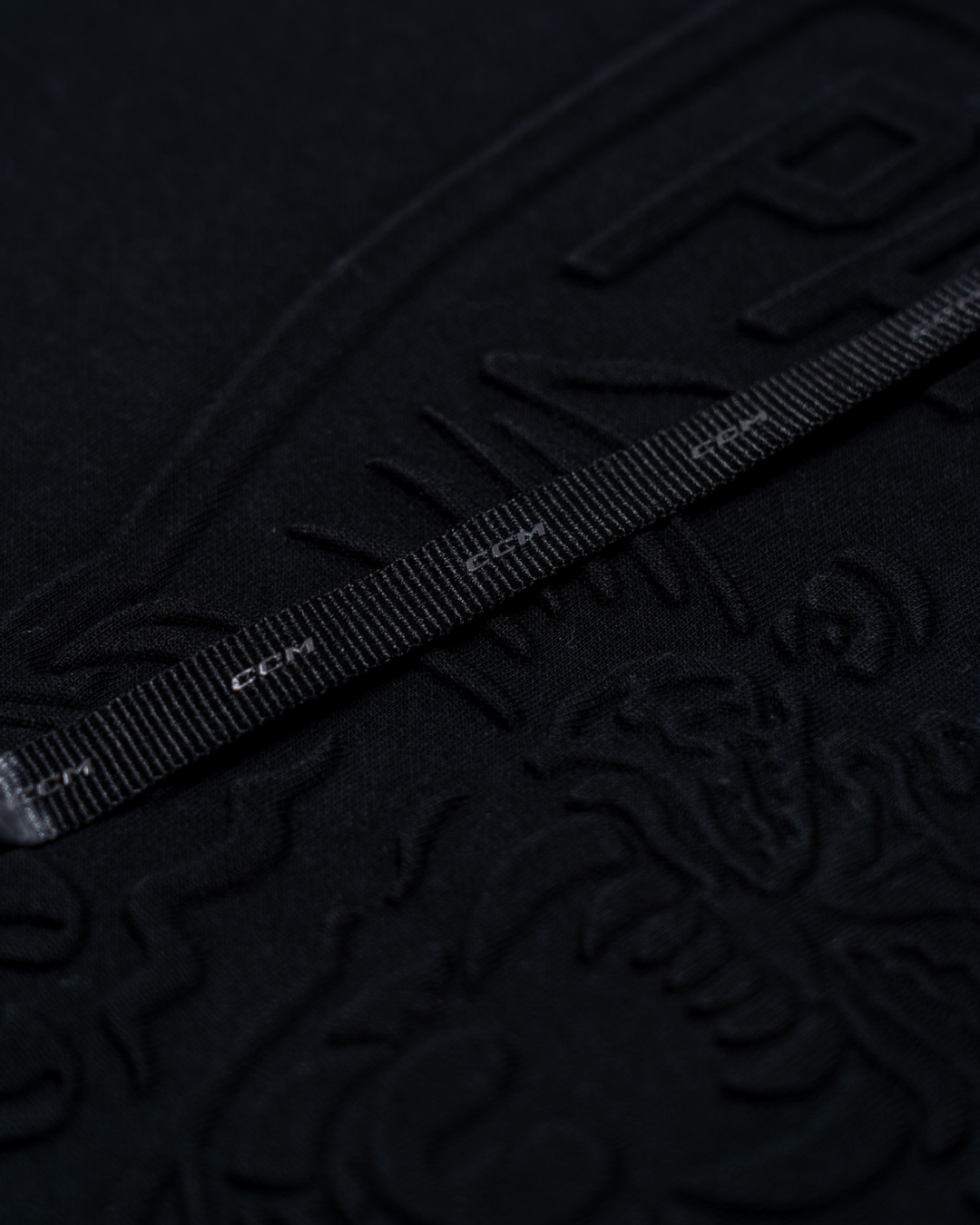 A close-up of the Panthersland Nottingham Panthers 25-26 CCM Black Embossed Hoodie shows its textured black surface with embossed patterns and text, plus a dragon-like design. A black measuring tape with white markings lays across the hoodie.