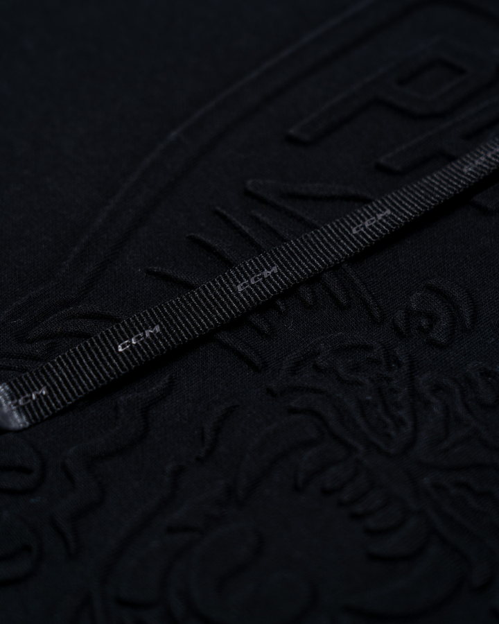 A close-up of the Panthersland Nottingham Panthers 25-26 CCM Black Embossed Hoodie shows its textured black surface with embossed patterns and text, plus a dragon-like design. A black measuring tape with white markings lays across the hoodie.