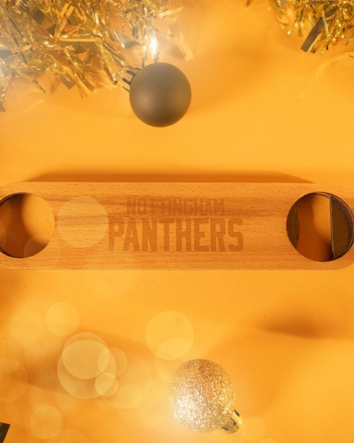 A Wooden Bottle Opener by The Nottingham Panthers sits on a yellow surface, accented with sparkling gold tinsel and Christmas ornaments for a festive holiday vibe.