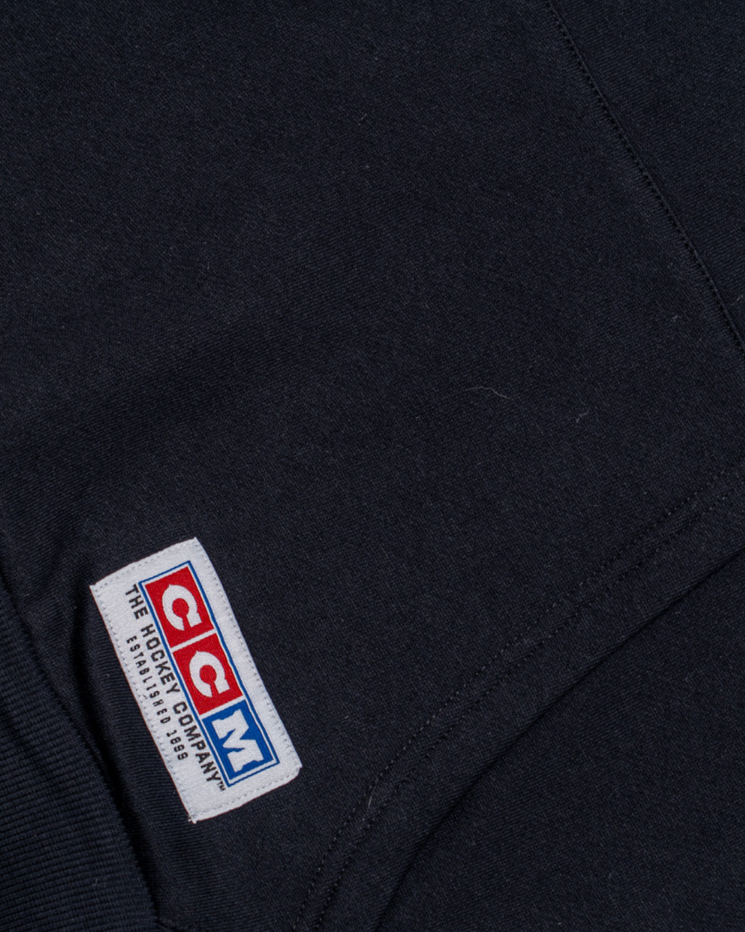 Close-up of the Nottingham Panthers 25-26 CCM Black Pullover Hoodie from Panthersland, showing a sewn rectangular patch with the CCM logo in red, white, and blue, plus "The Hockey Company Since 1899.