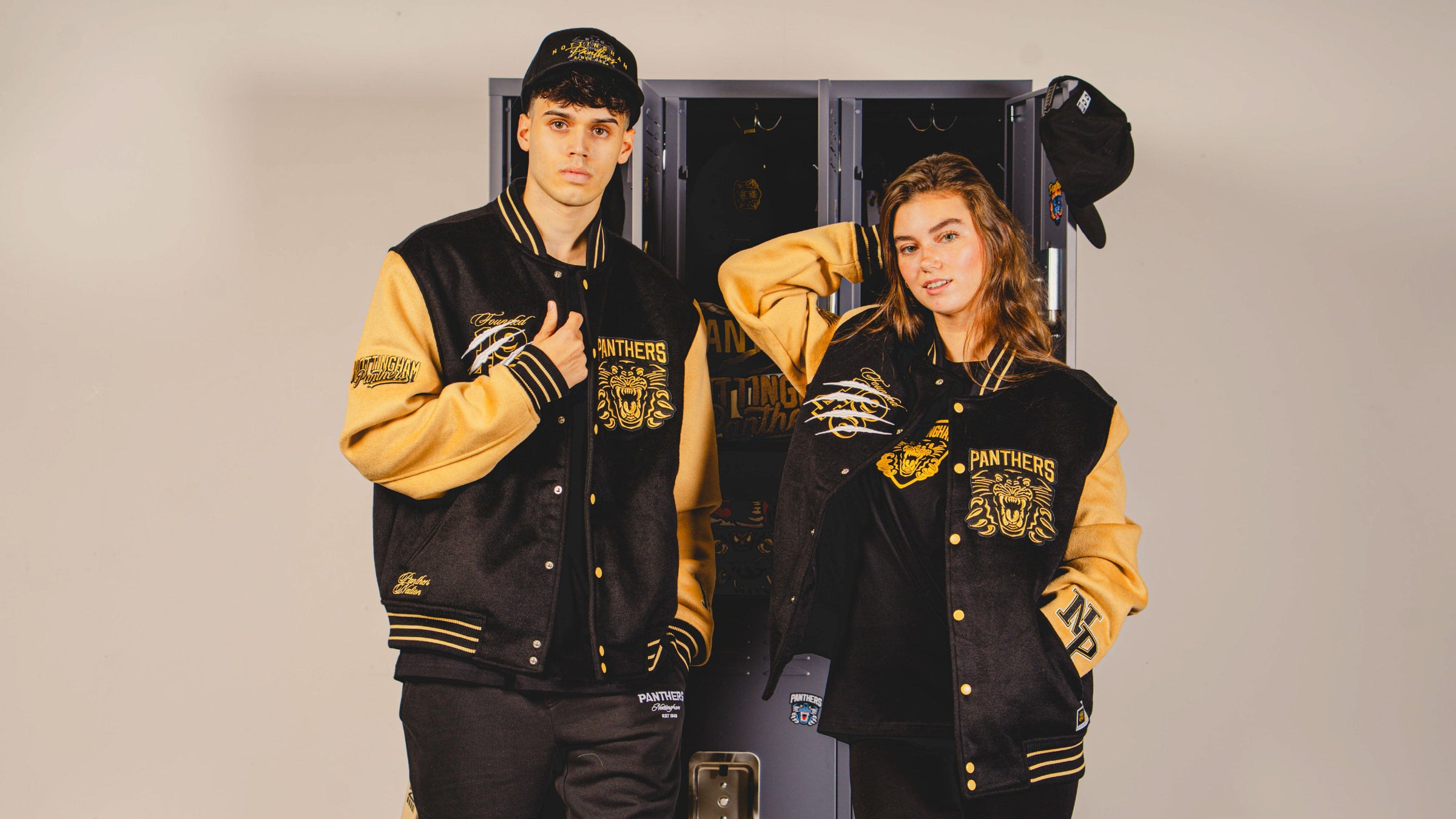 Two young adults pose confidently in black and gold varsity jackets and matching hats, standing in front of a locker with a Panthers sports team theme.
