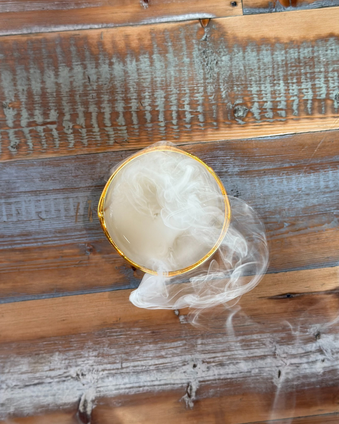 A round glass with smoky vapor, created by the Panthersland Nottingham Panthers Whiskey Smoker Kit, sits on a rustic wooden table with distressed paint.