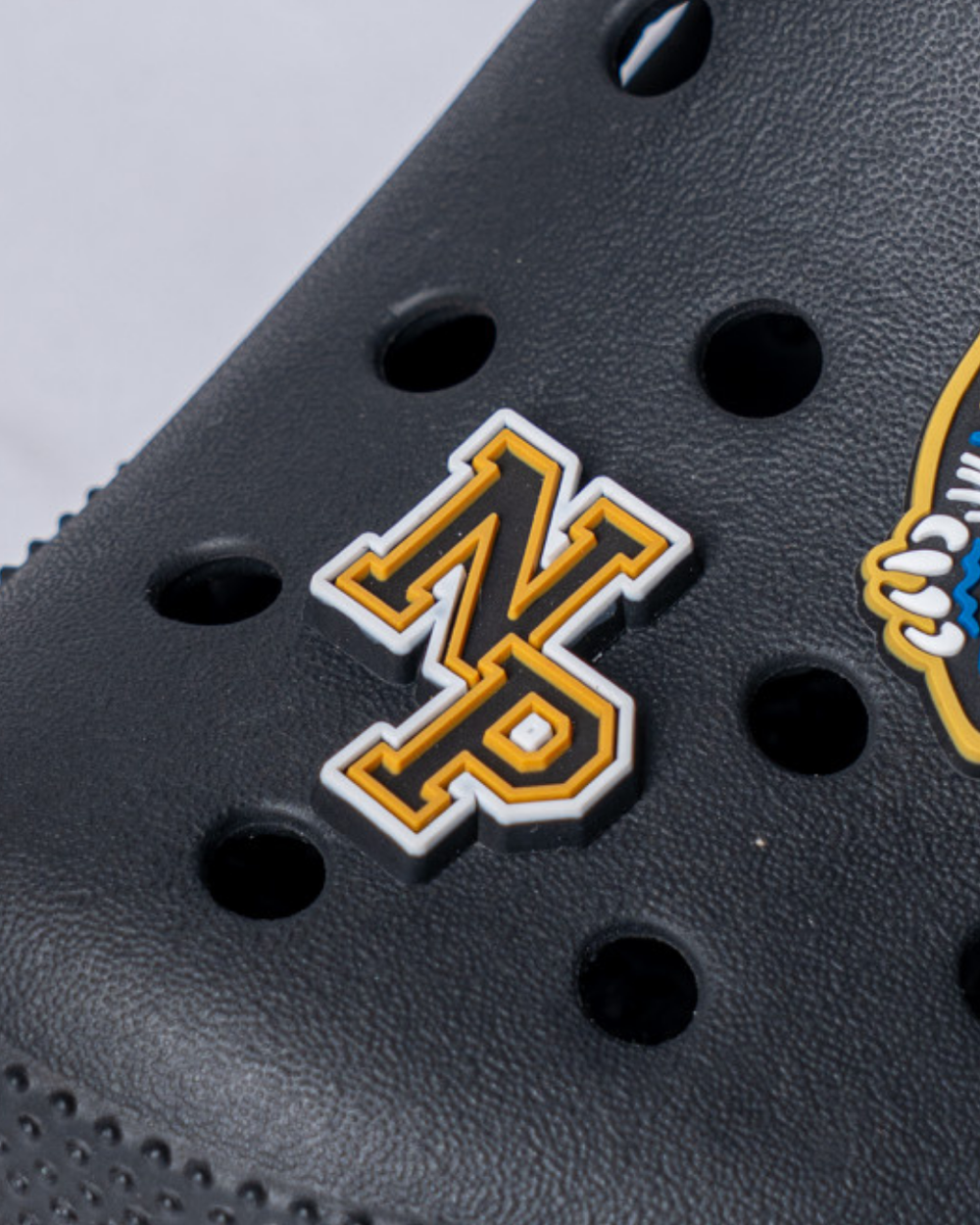 A close-up of a black shoe with ventilation holes, featuring the Nottingham Panthers 25-26 NP Croc Charm by Panthersland in yellow and white attached.