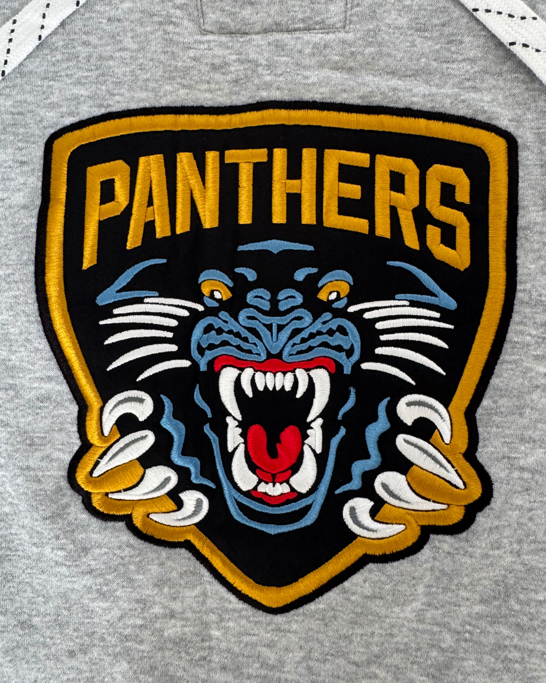 Add bold style to your 26-27 Grey Lace Crewneck from Panthersland with an embroidered patch featuring a snarling panther—yellow eyes and claws—set on a black and gold shield with “PANTHERS” above the animal, all sewn onto a gray fabric background. Perfect for pairing with pieces that showcase a twill crest logo or showing off your Panthers pride.