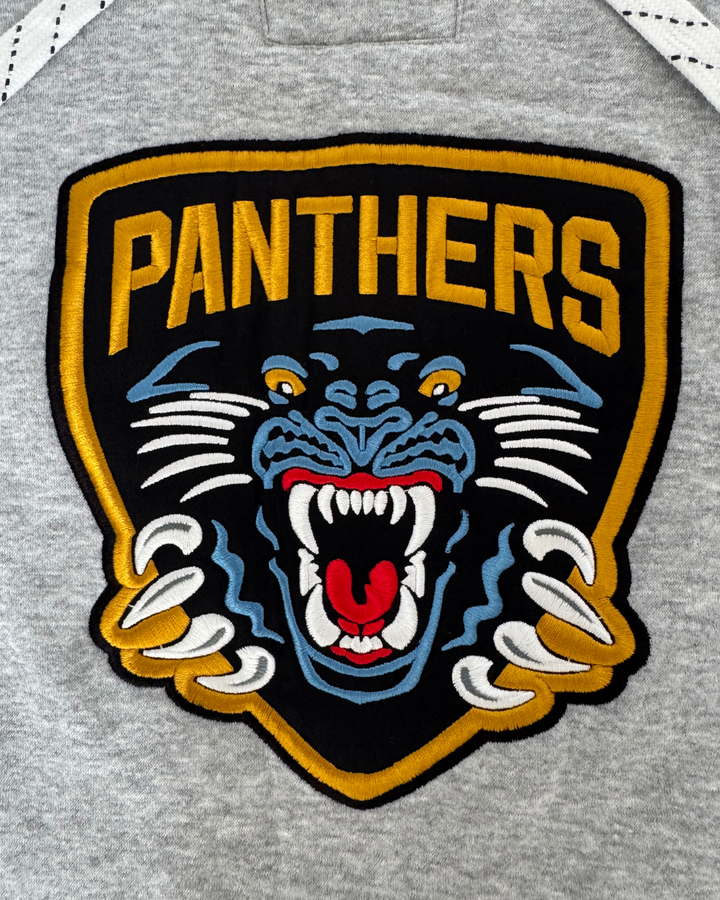 Add bold style to your 26-27 Grey Lace Crewneck from Panthersland with an embroidered patch featuring a snarling panther—yellow eyes and claws—set on a black and gold shield with “PANTHERS” above the animal, all sewn onto a gray fabric background. Perfect for pairing with pieces that showcase a twill crest logo or showing off your Panthers pride.