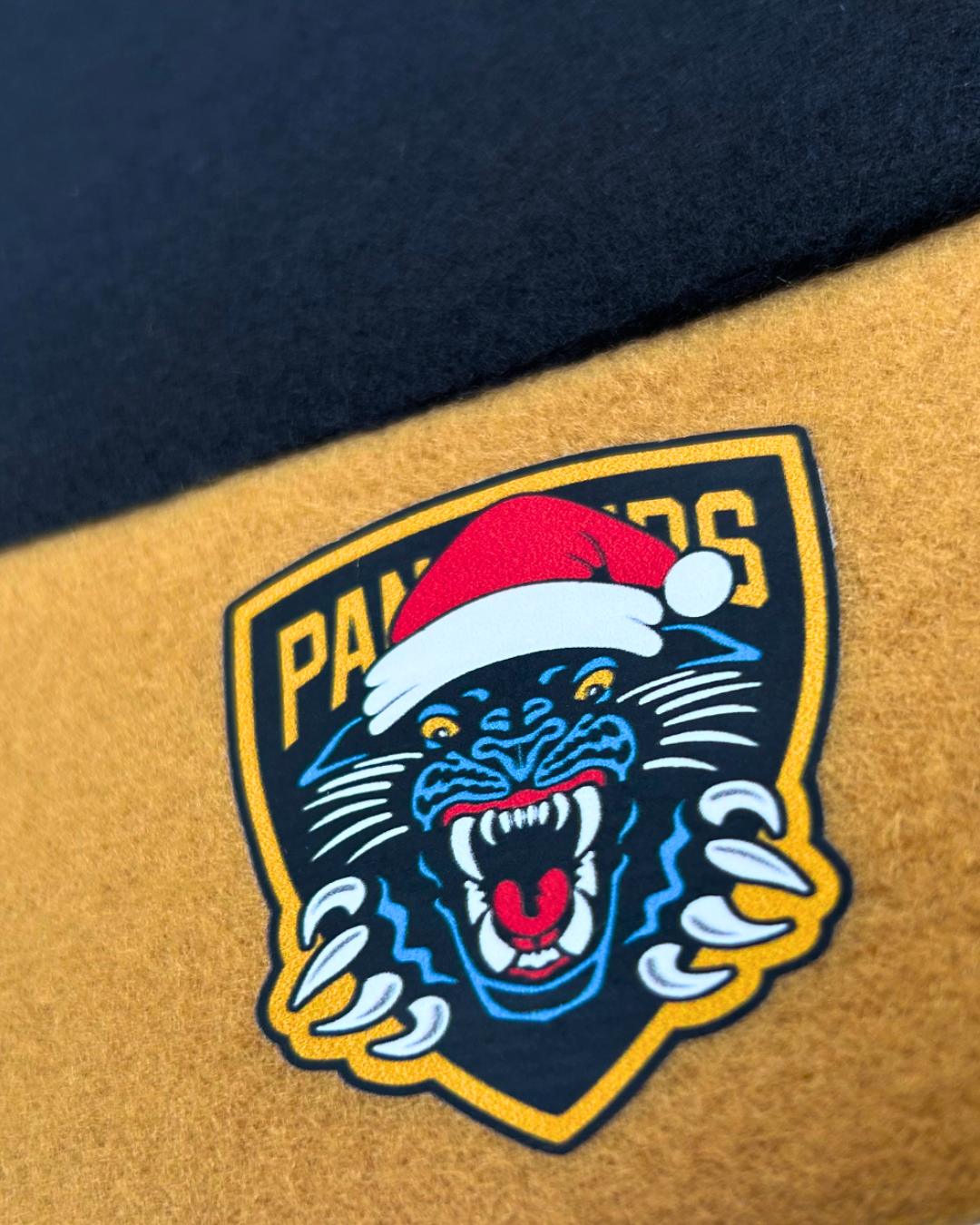 Celebrate the festive season in true Panthersland style with the Nottingham Panthers Santa Hat, featuring a snarling panther logo wearing a vibrant red Santa hat and “PANTHERS” displayed above—all printed on a striking yellow and black fabric background.