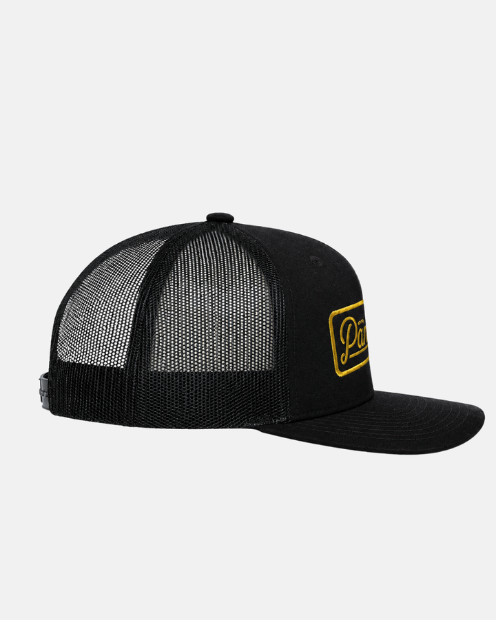 The Panthersland 26-27 Script Trucker Cap is a black mesh trucker hat with a flat brim, featuring a yellow embroidered Nottingham Panthers patch on the front. It has an adjustable snap closure at the mesh back, and the image is presented on a plain white background.