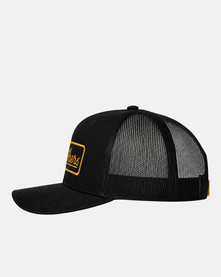 The 26-27 Script Trucker Cap by Panthersland features a black design with a curved brim, mesh back panels, and a yellow-bordered rectangular Nottingham Panthers patch on the front. The side view highlights its structured crown and adjustable snapback closure.