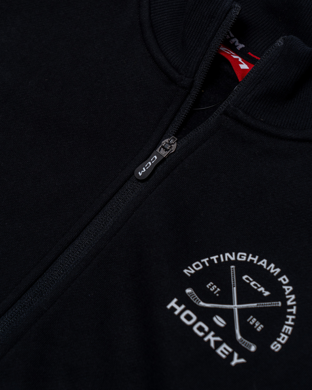 Close-up of a black Nottingham Panthers 25-26 CCM Track Jacket by Panthersland, featuring a white logo with crossed hockey sticks, "EST. 1946," and the CCM branding beneath the zipper.
