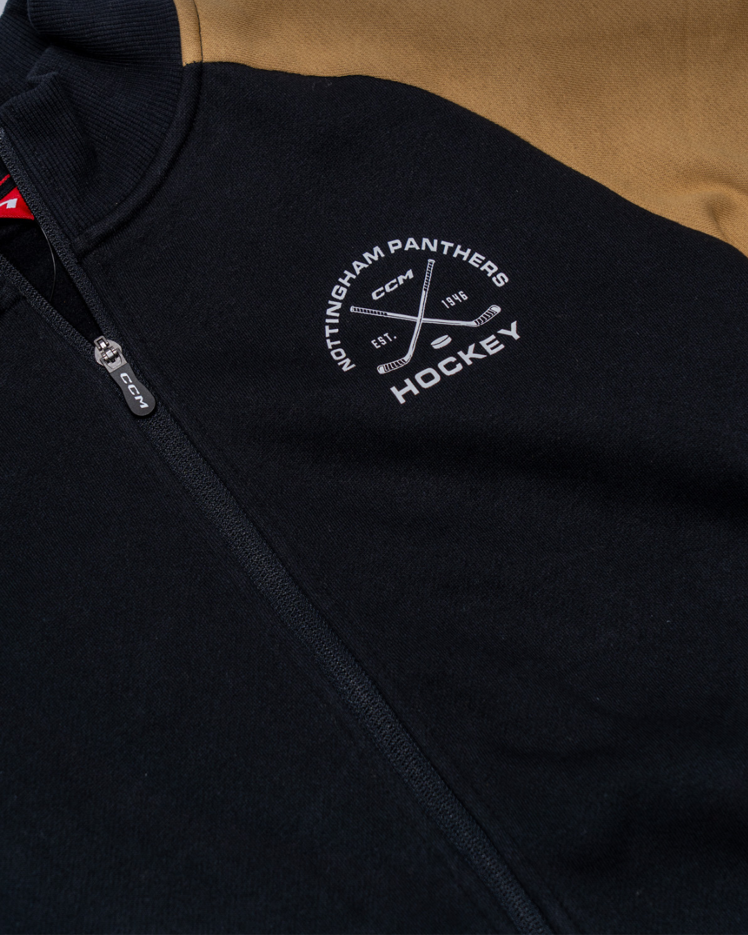 A close-up of the Panthersland Nottingham Panthers 25-26 CCM Track Jacket shows "Nottingham Panthers Hockey" and two white crossed hockey sticks embroidered on the black chest, with a contrasting tan sleeve.