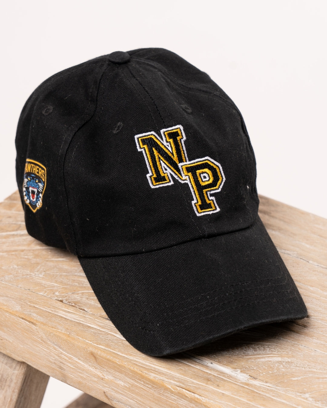 The Nottingham Panthers NP Logo Black Cap features bold yellow NP letters on the front and a vibrant Panthers patch on the side, shown here on a light wooden surface.
