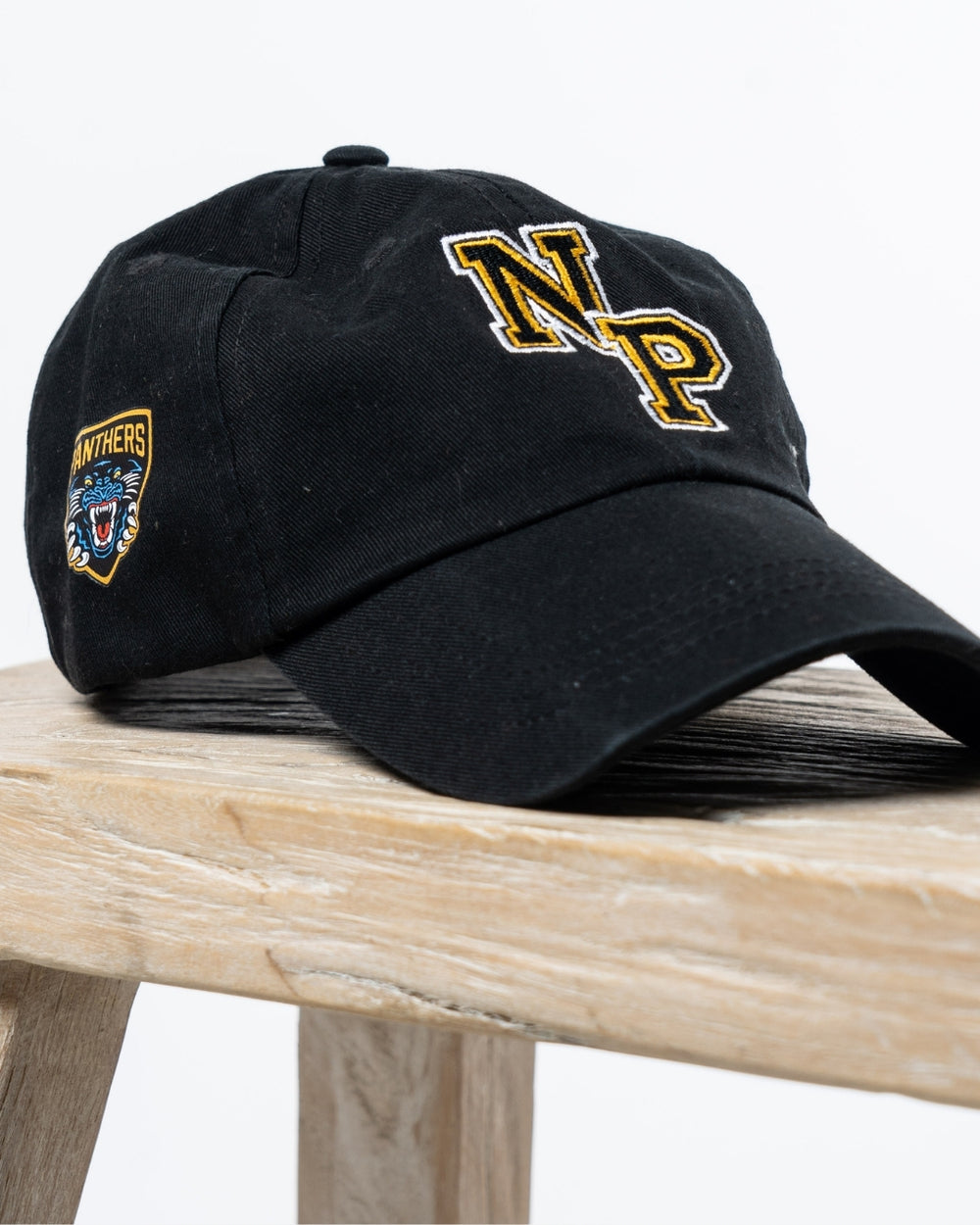 The NP Logo Black Cap by Nottingham Panthers, featuring gold and white NP letters on the front and a side panther Panthers patch, is stylishly displayed on a wooden surface.