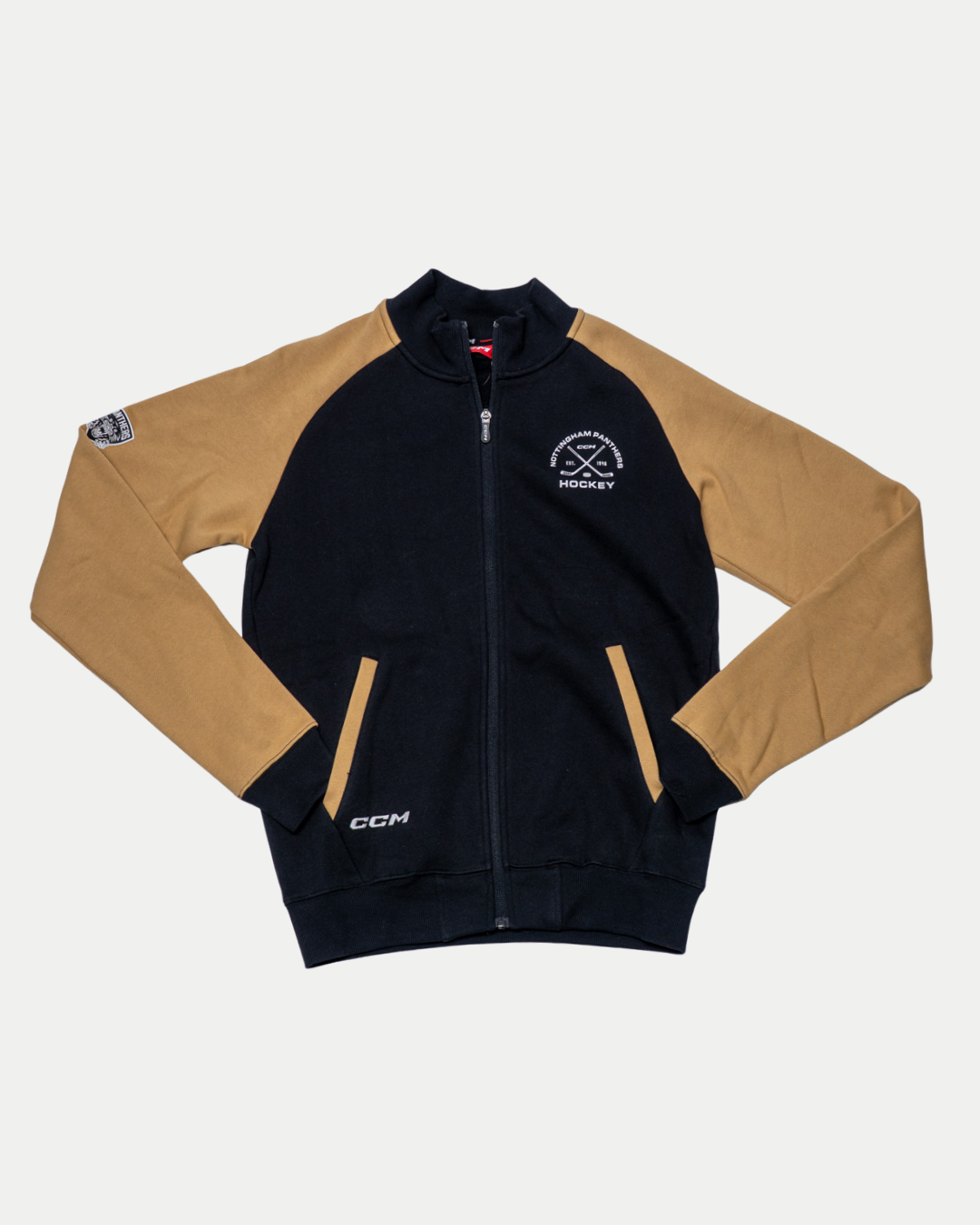 The Nottingham Panthers 25-26 CCM Track Jacket by Panthersland is a black and tan zip-up with tan sleeves, black body and collar, a crossed hockey stick HOCKEY logo on the chest, circular patch on the sleeve, and CCM branding near the hem.