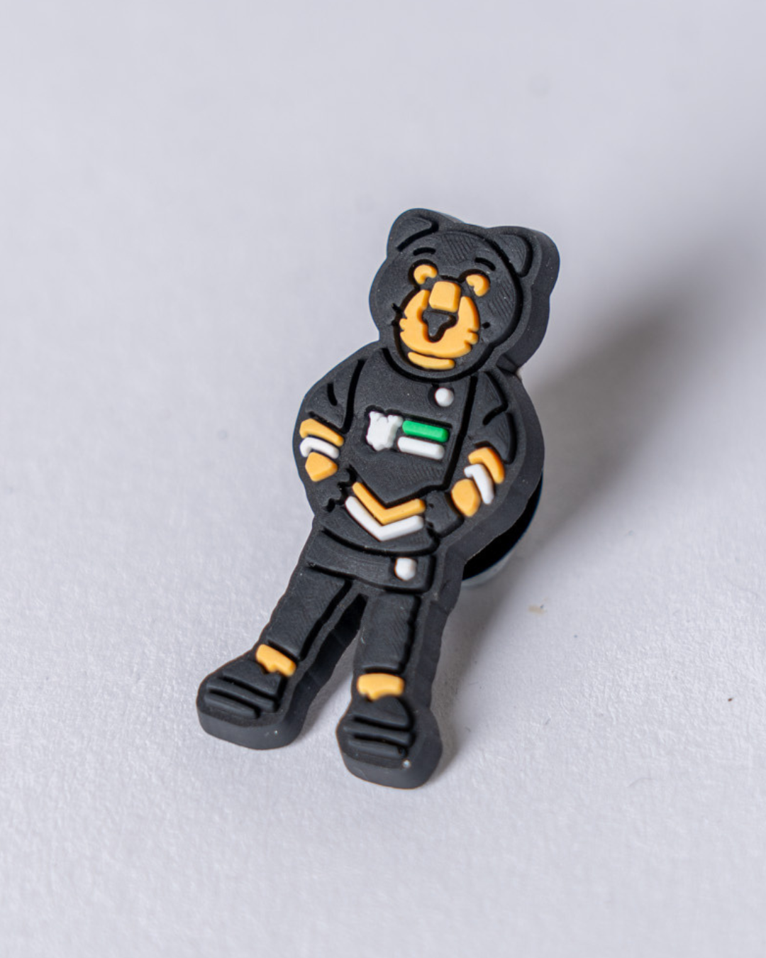 The Panthersland Paws Croc Charm is a rubber mascot pin shaped like a bear in a black astronaut suit with yellow accents and a flag, perfect for Crocs, shown on a light gray surface.