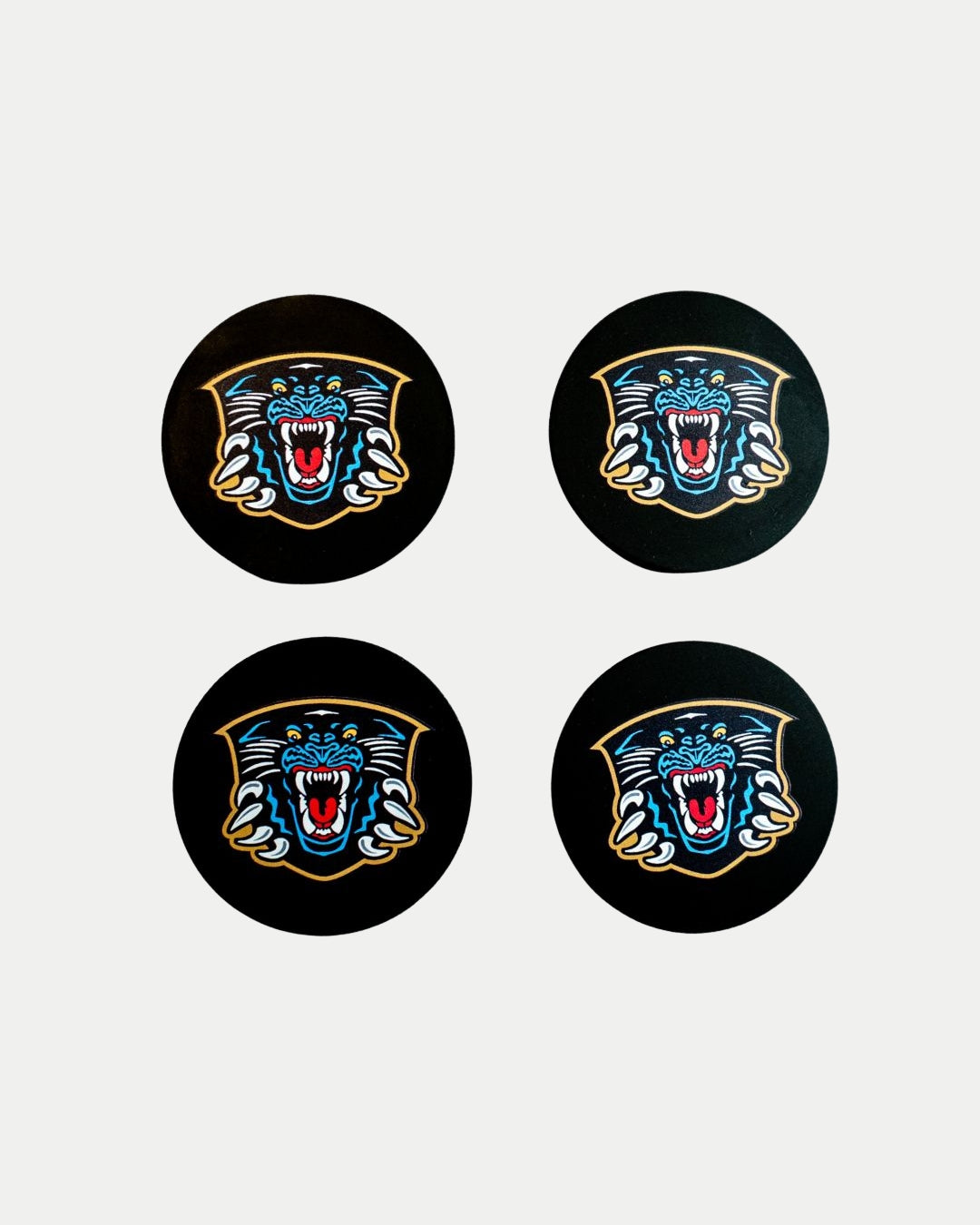 The Nottingham Panthers Puck Coaster Set by The Nottingham Panthers features four black coasters with a fierce panther head in blue and yellow, arranged neatly in a square on a white background.