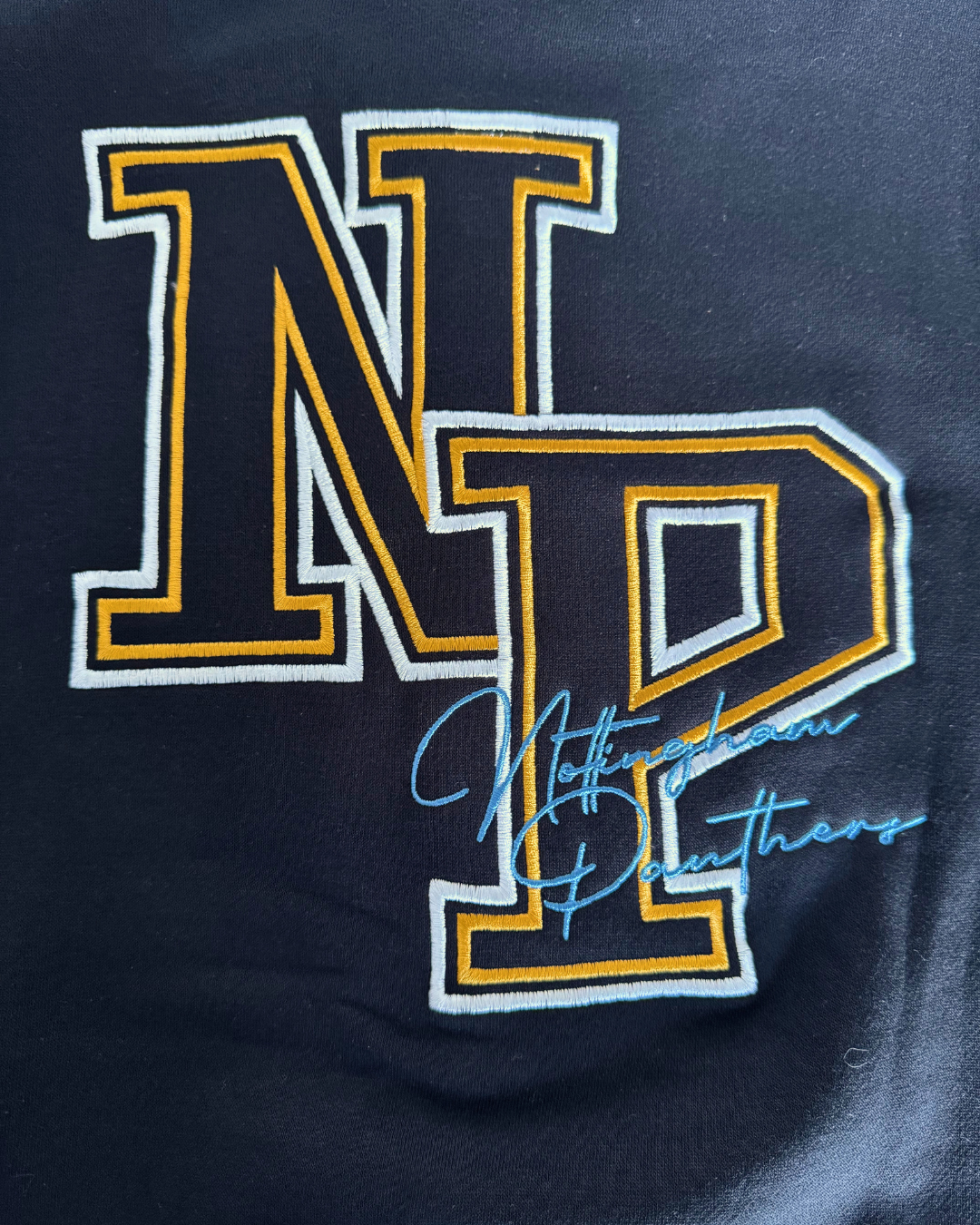 On a dark fabric background, the 26-27 NP Varsity Sweater by Panthersland features large yellow and white NP letters outlined in black in a classic varsity style, with “Nottingham Panthers” written below in light blue cursive.