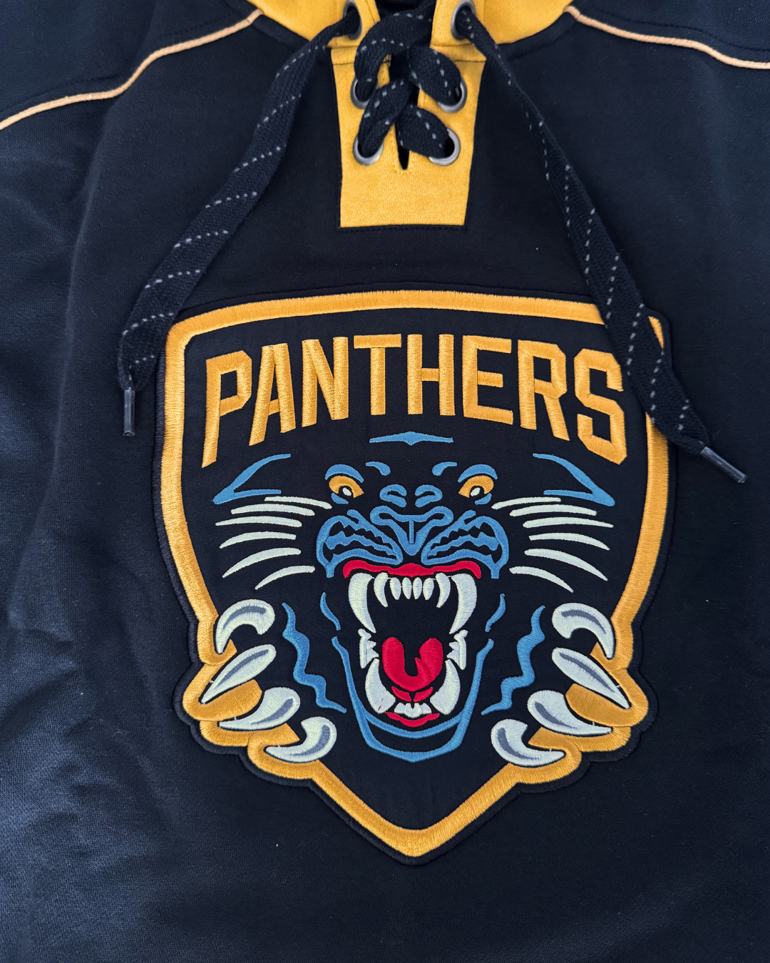 Close-up of the Panthersland 26-27 NP Lace Hoodie, a black hockey-style jersey featuring a fierce panther logo with the word “PANTHERS” above it. The design includes yellow accents and black laces near the collar, creating a bold sportswear look reminiscent of classic gold-stripe hoodies.