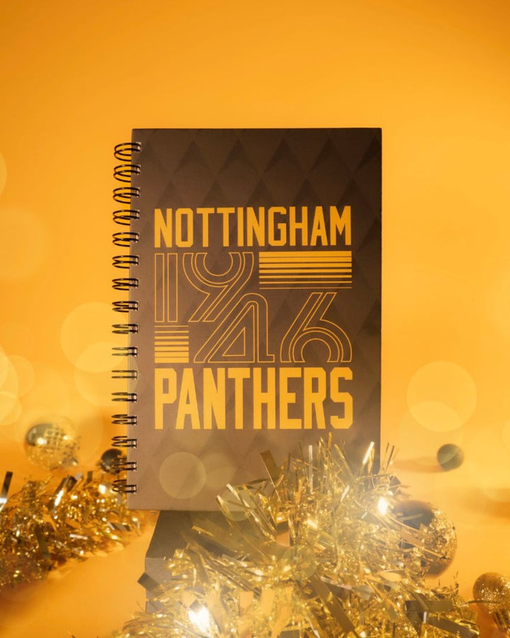 The A5 Crop Block Logo Notebook by The Nottingham Panthers features bold 1946 yellow text on the cover, surrounded by golden tinsel and festive lights, capturing team spirit against a warm yellow background.