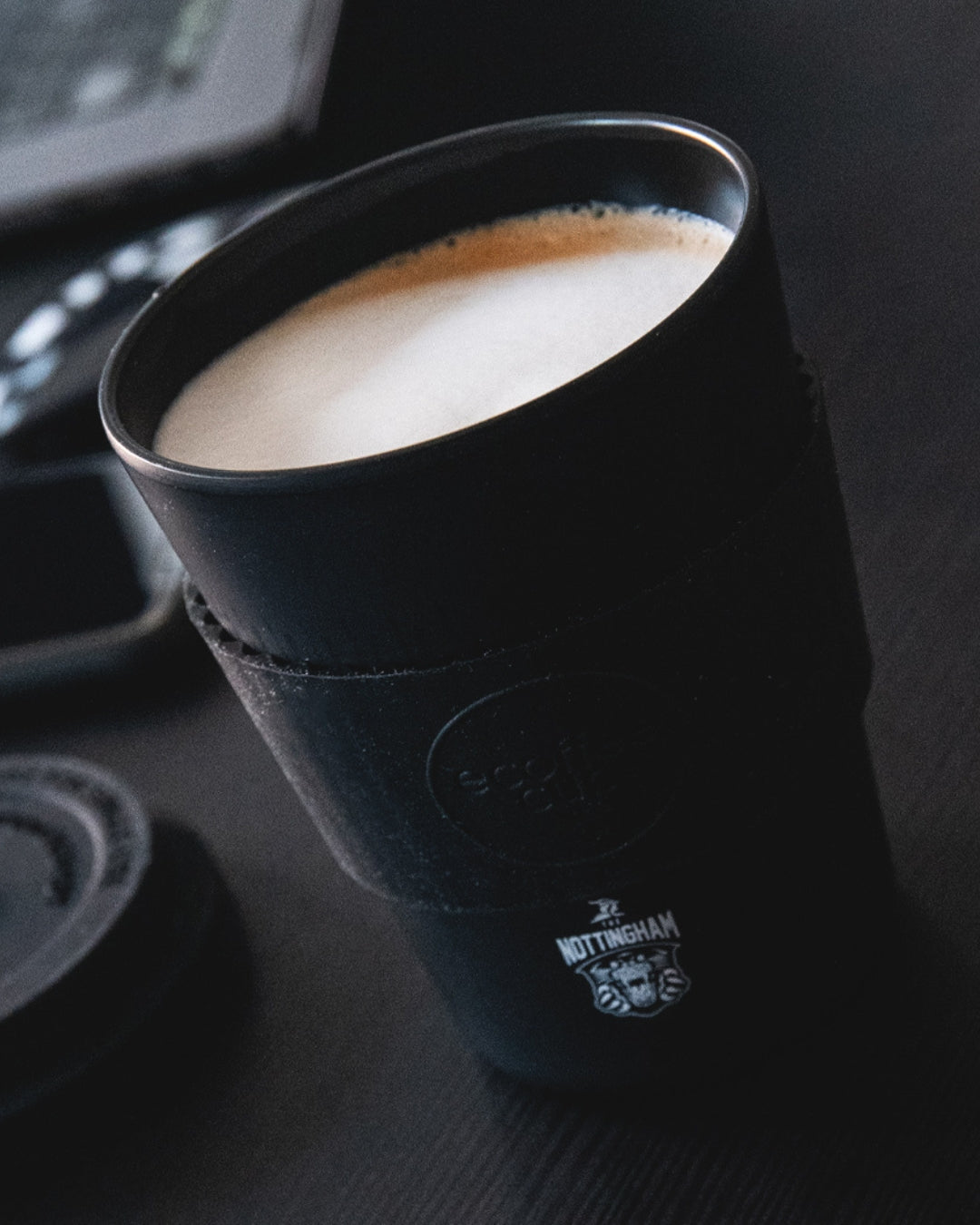 A Nottingham Panthers Reusable Cup sits on a dark surface, filled with a foamy drink. The cup features the teams lion logo, while its lid and an electronic device are blurred in the background.