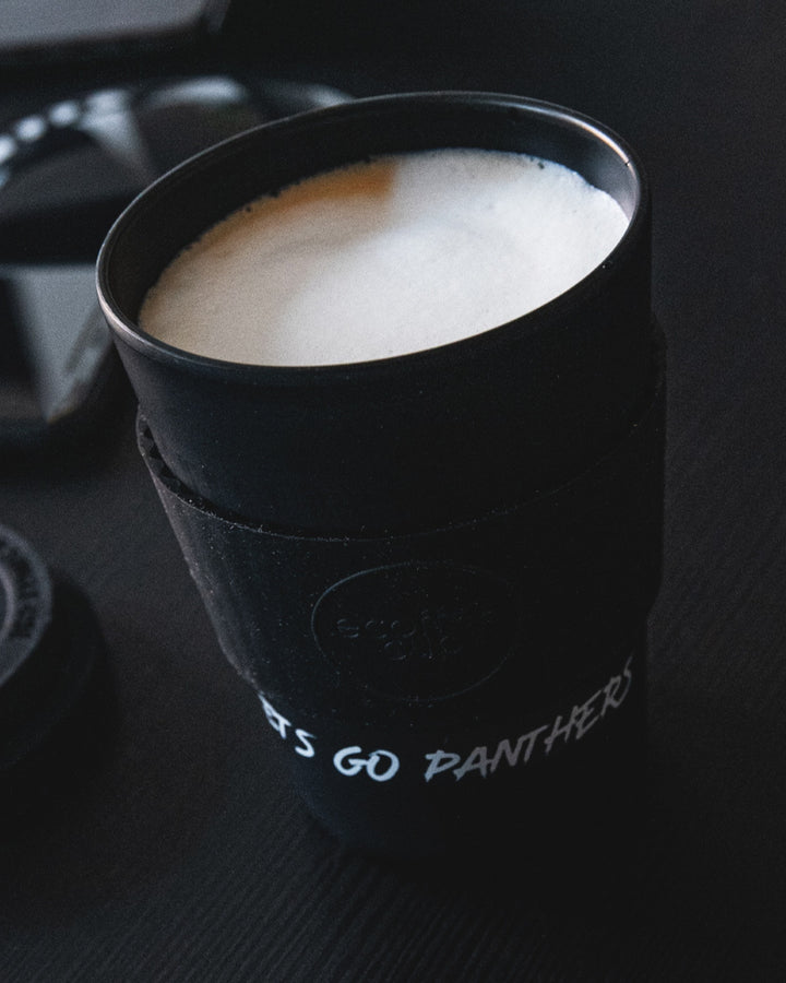 A Nottingham Panthers Reusable Cup, branded in black and eco-friendly, is filled with frothy latte. LET’S GO PANTHERS is partly visible on the sleeve as it rests on a dark surface.