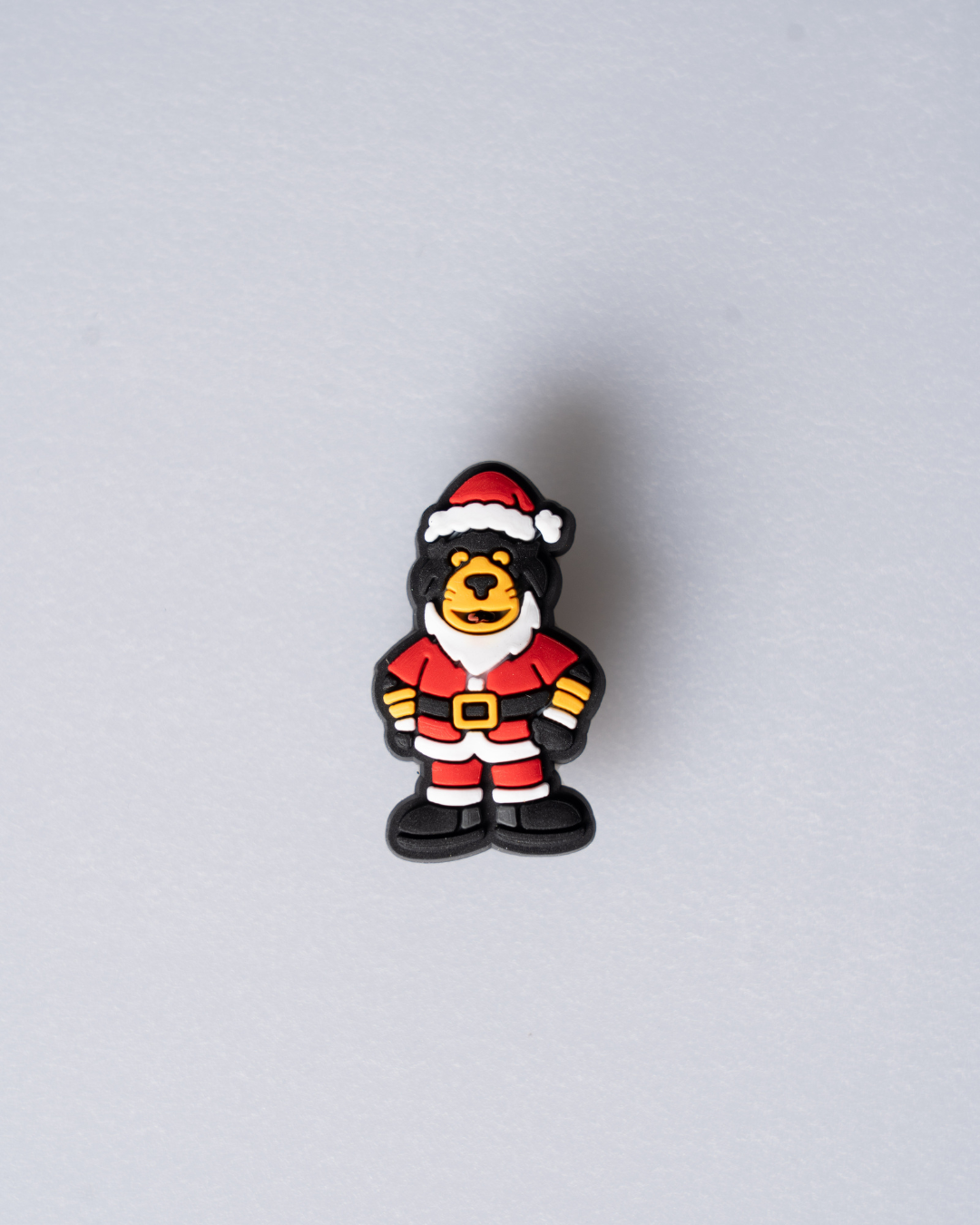 The Panthersland Santa Paws Croc Charm features a small cartoon bear in a Santa outfit—red hat, suit, black boots, and yellow belt—standing against a light gray background. Perfect for adding festive flair to your Crocs!.
