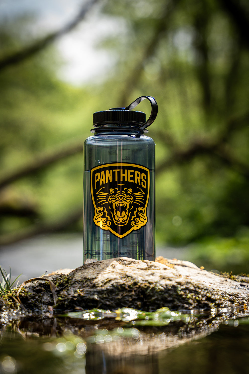 A Panthersland Panthers x Nalgene 1L Water Bottle with a black lid and a striking PANTHERS logo featuring a roaring panther stands on a rock by a stream in a lush, green forest. This wide-mouth bottle is perfect for keeping you hydrated during your outdoor adventures.