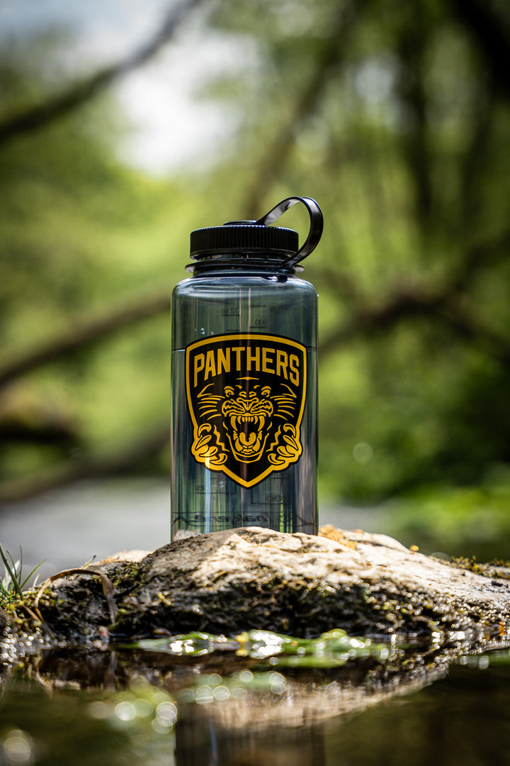 A Panthersland Panthers x Nalgene 1L Water Bottle with a black lid and a striking PANTHERS logo featuring a roaring panther stands on a rock by a stream in a lush, green forest. This wide-mouth bottle is perfect for keeping you hydrated during your outdoor adventures.