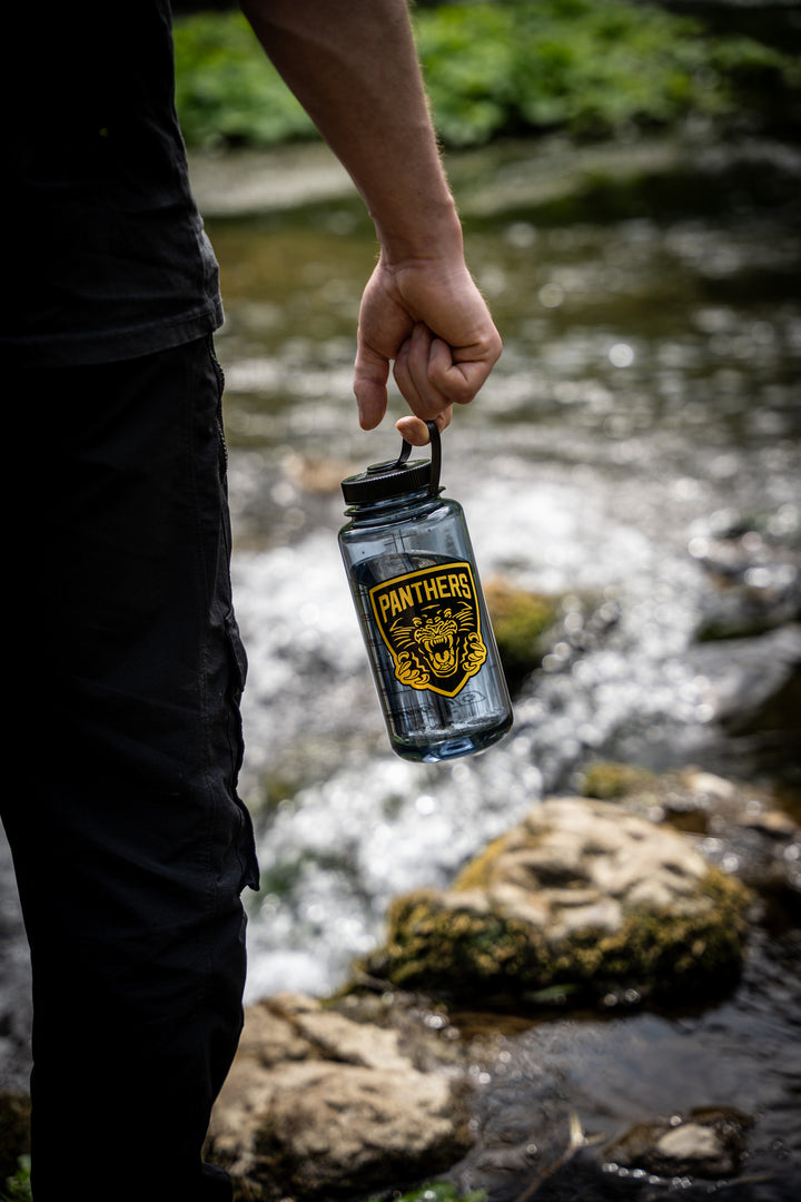 A person standing by a rocky stream holds the Panthers x Nalgene 1L Water Bottle from Panthersland, featuring a yellow logo with a tiger face. The wide mouth water bottle stands out against the lush greenery in the background.