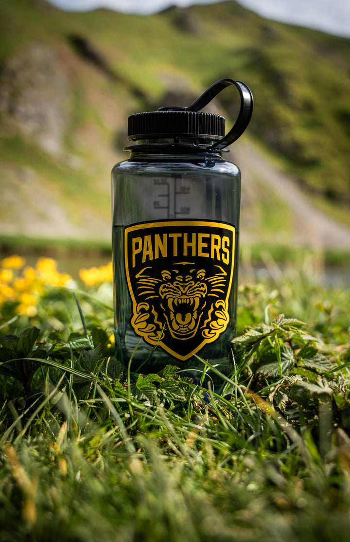A Panthersland Panthers x Nalgene 1L Water Bottle—a black, BPA-free bottle featuring a wide mouth and a vibrant yellow logo—rests on grass sprinkled with yellow flowers, set against mountains and a softly blurred natural backdrop. Ideal for outdoor adventures.
