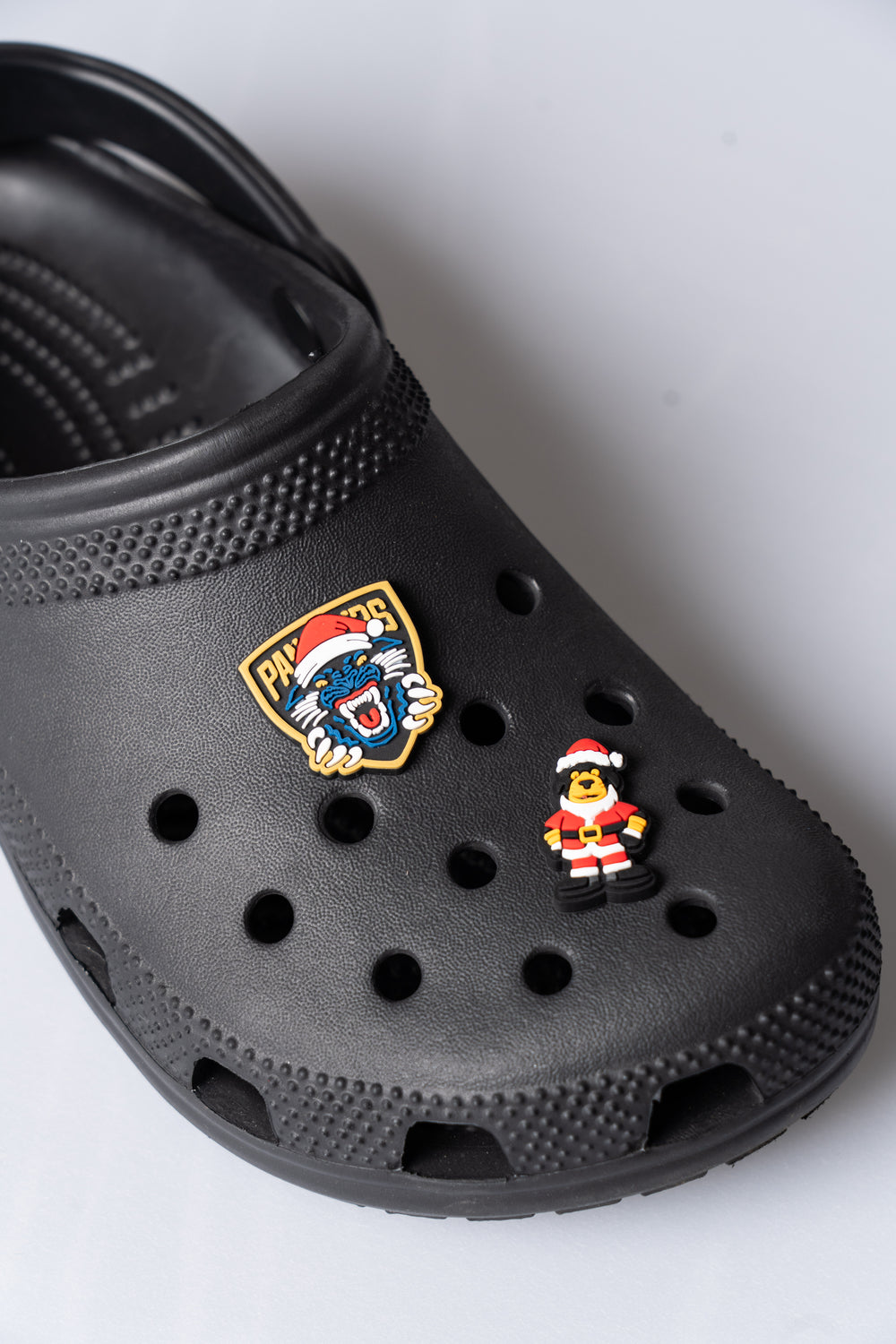 A close-up of a black clog sandal decorated with two pins: the Panthersland Nottingham Panthers Santa Crest Croc Charm featuring a lion head in a Santa hat, and another festive accessory—a small figure dressed as Santa Claus.