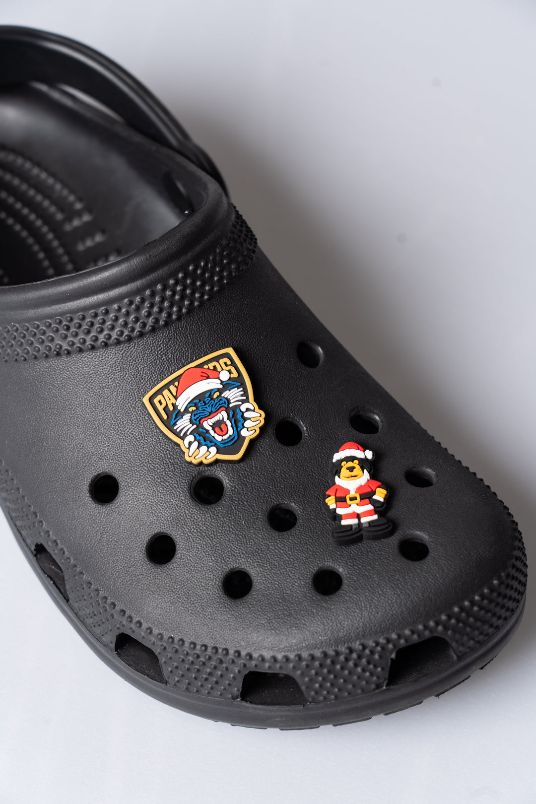 A close-up of a black clog sandal decorated with two pins: the Panthersland Nottingham Panthers Santa Crest Croc Charm featuring a lion head in a Santa hat, and another festive accessory—a small figure dressed as Santa Claus.