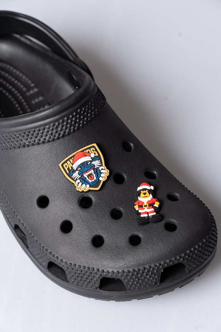A close-up of a black clog sandal decorated with two pins: the Panthersland Nottingham Panthers Santa Crest Croc Charm featuring a lion head in a Santa hat, and another festive accessory—a small figure dressed as Santa Claus.