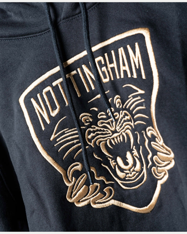 Close-up of the Golden Twill Hockey Club Hoodie Junior by The Nottingham Panthers, featuring NOTTINGHAM in bold above an embroidered roaring panther face with outstretched claws; embroidery uses beige and white thread.