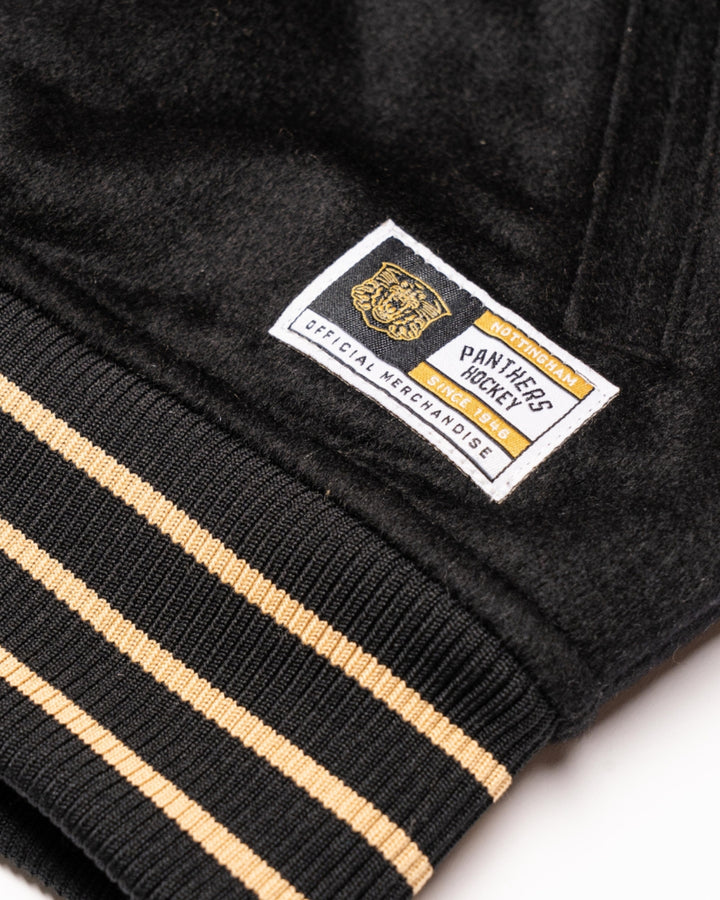 Close-up of the Nottingham Panthers Varsity Jacket Black/Gold 25-26 by Panthersland, highlighting beige and black striped cuffs and a stitched patch reading Panthers PS Hockey Society Official Merchandise with a bold panther design.