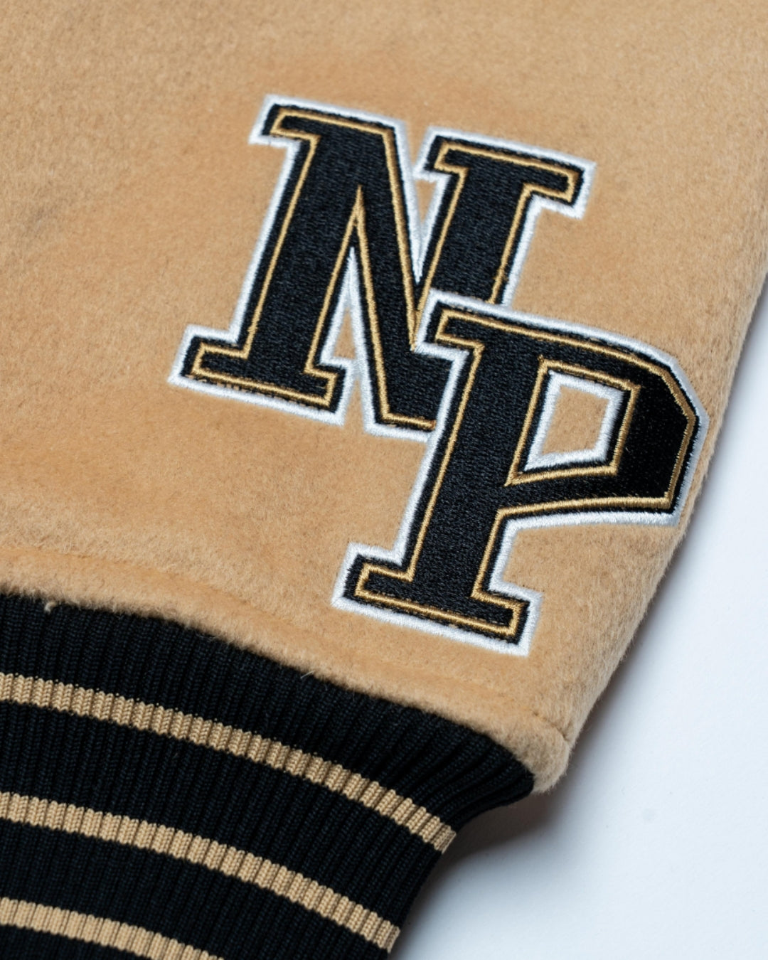 Close-up of the Nottingham Panthers Varsity Jacket Black/Gold 25-26 by Panthersland, showing tan fabric with black and gold varsity-style “N” and “P” embroidery beside a black ribbed cuff with tan stripes.