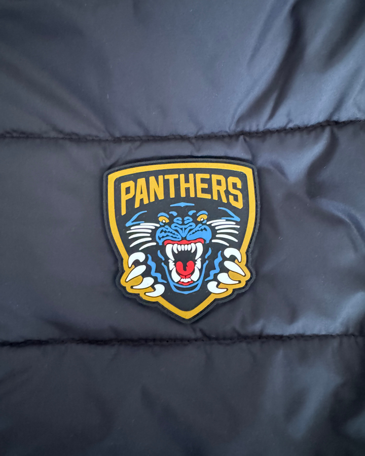 A silicone crest featuring a roaring blue panther inside a yellow shield with bold yellow “PANTHERS” lettering sits prominently on the 26-27 Padded Crest Jacket by Panthersland, adding a striking detail to this classic Nottingham Panthers outerwear.