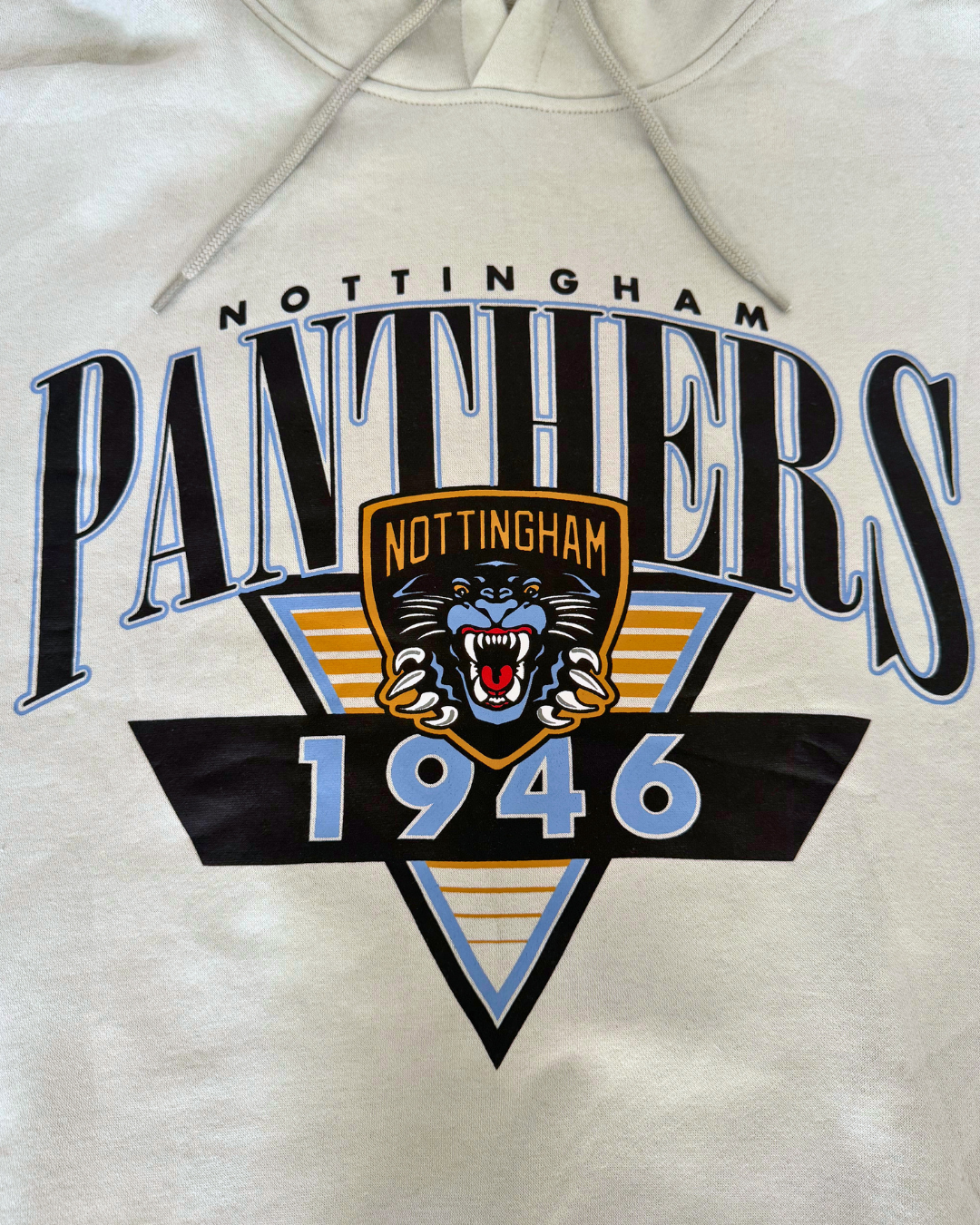 The Panthersland 26-27 Vintage Hoodie is a beige sweatshirt showcasing a retro Nottingham Panthers 1946 design, highlighted by a central roaring panther crest logo, bold blue and gold accents, and an eye-catching triangular background pattern.